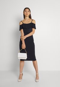 Vestido azul-marinho com decote ombro a ombro, detalhamento em babados, design ajustado e corte até ao joelho. Acessórios incluem uma pequena mala branca.