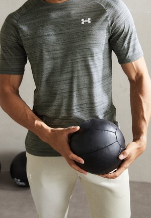 Personne portant un t-shirt de sport gris et un pantalon blanc tenant un ballon médicinal noir au niveau de la taille à l’intérieur.