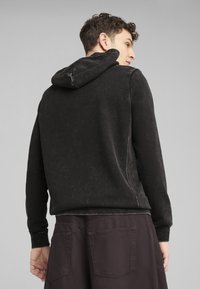 Felpa nera con finitura testurizzata, dotata di cappuccio con cordino, maniche lunghe e dettagli delle cuciture visibili. Il modello indossa pantaloni scuri e larghi.
