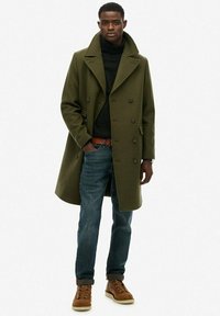 Abrigo de doble botonadura en verde oliva sobre un suéter negro de cuello alto, combinado con jeans azules y botas marrones con cordones. Tejido texturizado, diseño a la altura de la rodilla.