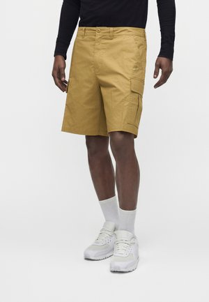 Homme portant un short cargo beige, des chaussettes blanches et des baskets blanches, debout devant un fond clair uni.