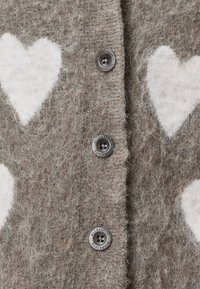 Gros plan d'un tissu tricoté marron duveteux avec des motifs de cœur blancs et des boutons argentés gravés avec "ALLSAINTS."