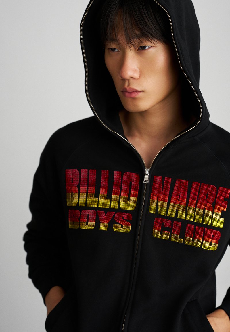 Crna zip-up dukserica s kapuljačom. Sadrži hrabre, višebojne natpise "BILLIONAIRE BOYS CLUB" u crvenoj i žutoj boji. Mekani materijal, opušteni kroj.