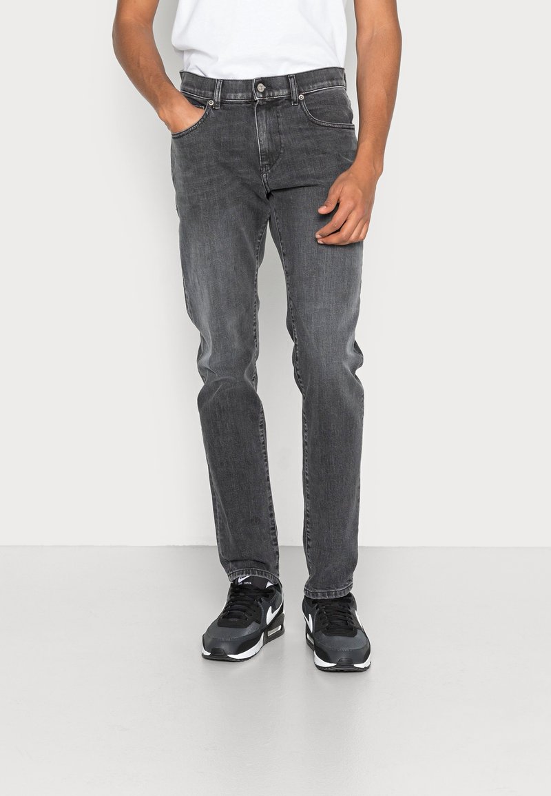 Jeans de mezclilla gris con un ajuste slim, diseño de cinco bolsillos y un ligero desgaste. Llevados con zapatos deportivos negros, de pie sobre un fondo neutro.