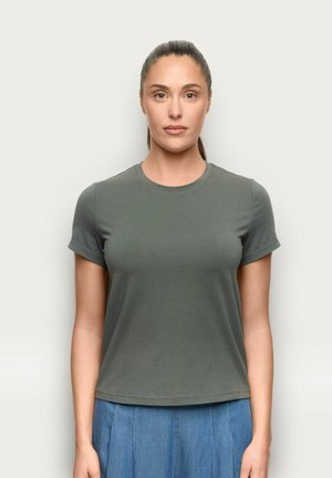 Kobieta z włosami zaczesanymi do tyłu, ubrana w szarą koszulkę z krótkim rękawem i niebieską plisowaną spódnicę, stojąca na jednolitym jasnym tle.