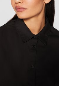 Black button-up shirt featuring a classic collar, smooth texture, and five black buttons along the front for closure.