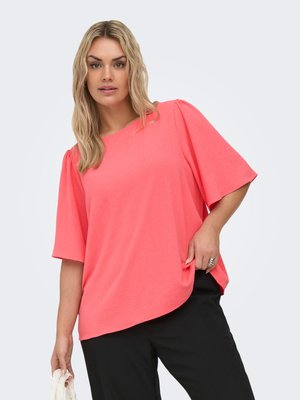 Femeie blondă cu valuri lejere, purtând o bluză coral cu mâneci scurte și pantaloni negri, ținând o țesătură albă în mâna stângă.