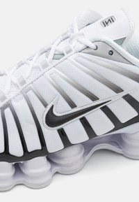 Chaussure de course Nike avec une tige en mesh blanche avec des accents noirs, des panneaux latéraux gris audacieux et des éléments de coussinage arrondis uniques.
