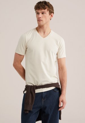 Fiatal férfi, aki krémszínű V-nyakú pólót és kék farmert visel, sötétbarna pulóvert kötött a derekára, semleges háttér előtt áll.