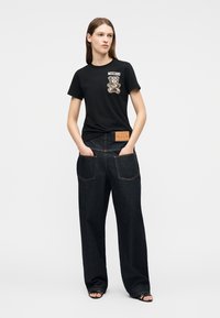 Mujer joven con una camiseta negra de Moschino con estampado de osito de peluche y vaqueros oscuros de pierna ancha, de pie con las manos en los bolsillos.