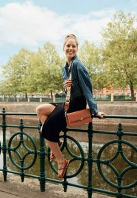 Hnědá kožená crossbody taška s texturovaným popruhem, modrá džínová bunda, černá sukně a hnědé sandály, umístěné na zeleném zábradlí u vody.