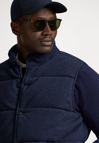 Granatowa pikowana kamizelka z teksturowanej tkaniny, z zapięciem na guziki i detalem z logo na piersi. Połączona z granatowym swetrem i czapką.