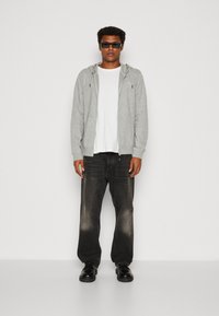 Gray zip-up hoodie over a white t-shirt, paired with faded black jeans and black chunky shoes. Accessories include dark sunglasses.