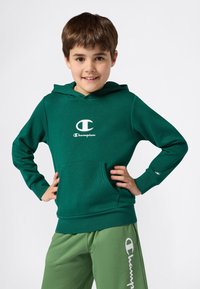 Ragazzo in felpa verde Champion e pantaloni della tuta verdi, in piedi con le mani sui fianchi, sorridente su uno sfondo bianco uniforme.
