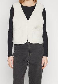 Ljusgrå fleeceväst med dragkedja, som har två framfickor och en åtsittande design, lager på en svart långärmad tröja.