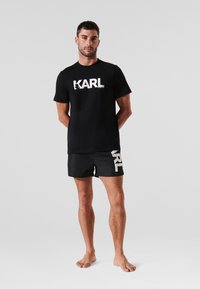 Crna pamučna majica s velikim bijelim logotipom "KARL" na prednjoj strani, u kombinaciji s crnim šortsem koji ima sličan logotip, minimalistički dizajn i kratke rukave.