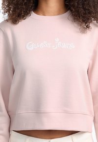 Light pink cropped sweatshirt made of soft fabric, featuring white embroidered "Guess Jeans" logo centered on the chest. Ribbed collar and hem.