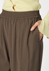 Brune viddebukser med elastisk liv, som har sidelommer og et glatt, lett teksturert stoff.
