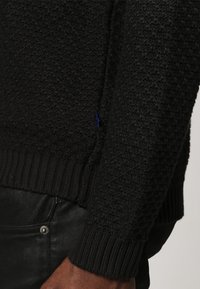 Maglione nero testé con un motivo a diamante, orlo a costine e un dettaglio con etichetta blu sul lato. Il tessuto sembra lavorato a maglia e spesso.