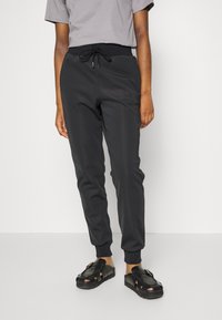 Black jogger pants with a fitted ankle cuff, elastic waistband with drawstring, side pockets, and smooth texture. Worn with black sandals.