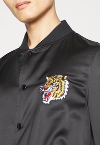 Homme portant une veste en satin noire avec des boutons noirs et une tête de tigre colorée brodée sur le côté gauche de la poitrine.