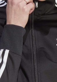 Chaqueta negra con cremallera y rayas blancas en las mangas. Cuenta con cierre de cremallera y puños acanalados. Textura de tela suave y etiqueta de marca visible.