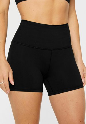 Woman wearing high-waist black athletic shorts and a black sports bra, showing midriff and upper thighs against a plain background.