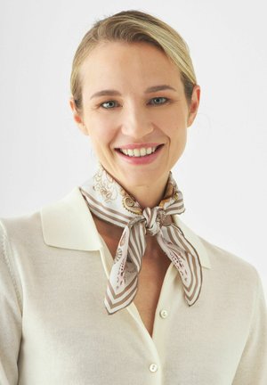 Lächelnde Frau mit blonden Haaren trägt eine cremefarbene, geknöpfte Bluse und einen gemusterten Schal, der um ihren Hals gebunden ist, vor einem einfarbigen Hintergrund.