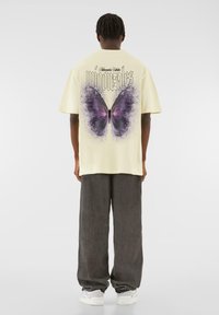Light yellow oversized t-shirt featuring a large purple butterfly graphic and text on the back, paired with loose, gray trousers and white sneakers.