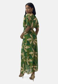 Maxikleid aus einem grünen Stoff mit einem botanischen Druck, der große Blätter in verschiedenen Grüntönen und Beige zeigt. Kurze Ärmel und elastischer Taillendetail.