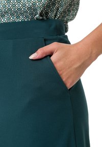 Smaragdově zelená tužková sukně s hladkou texturou, která má boční kapsu a vysoký pas. Složená s vzorovanou blůzkou nahoře.