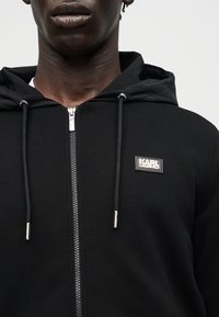 Záber zblízka na osobu, ktorá má na sebe čiernu mikinu so zipsom, kapucňou, sťahovacími šnúrkami a malou nášivkou s logom „Karl Lagerfeld“ na hrudi.