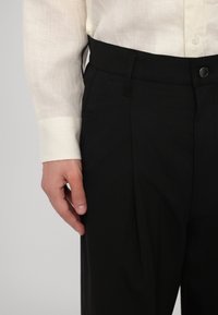 Pantaloni neri con una texture liscia, dotati di chiusura con bottone e tasche laterali, abbinati a una camicia bianca leggera e trasparente.