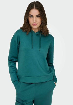 Jeune femme aux cheveux châtain mi-longs portant un sweat à capuche turquoise et un pantalon assorti, les mains dans les poches, sur un fond clair uni.