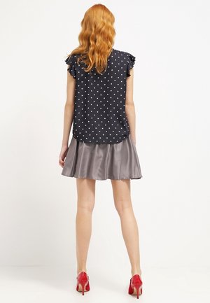 Granatowa bluzka w groszki z falbaniastymi rękawami, zestawiona z szarą satynową rozkloszowaną spódnicą, noszona z czerwonymi butami na wysokim obcasie.