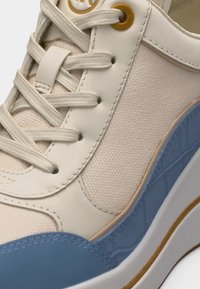 Κοντινή λήψη μπεζ και μπλε αθλητικού παπουτσιού με υφασμάτινες και δερμάτινες υφές, λευκά κορδόνια και χρυσή λεπτομέρεια με οπές κοντά στο επάνω μέρος.