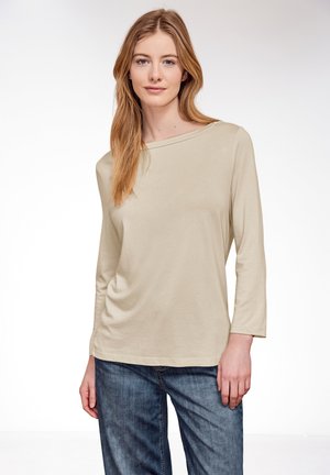 Junge Frau mit hellbraunen Haaren, die ein beiges Langarmshirt und blaue Jeans trägt, vor einem schlichten weißen Hintergrund stehend.