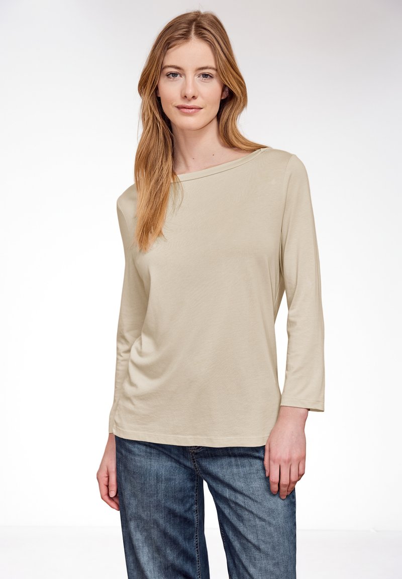 Jeune femme aux cheveux châtain clair portant un haut beige à manches longues et un jean bleu, debout devant un fond blanc uni.