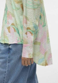 Blusa leggera a fantasia floreale in tonalità pastello. Presenta un tessuto texturizzato, maniche lunghe e polsini con bottoni, abbinata a jeans.