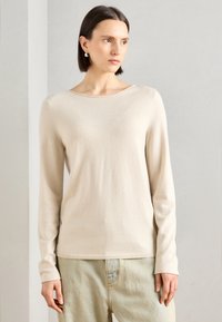 Lys beige langermet genser med en jevn tekstur og rund hals, kombinert med lyse jeans. Enkelt design uten mønstre eller dekorasjoner.