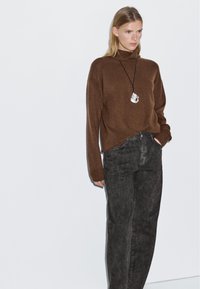 Maglione a collo alto marrone in tessuto morbido, abbinato a jeans neri a vita alta. Una collana con pendente in argento aggiunge un tocco di dettaglio. Look semplice e casual.