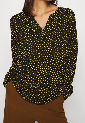 Blouse noire à manches longues avec un petit imprimé floral jaune et blanc, portée avec un pantalon marron, mains dans les poches.