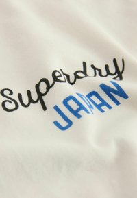 Balts audums ar "Superdry" melnā kursīvā un "JAPAN" zilās blokvēstulēs. Tekstūra šķiet maiga un gluda.