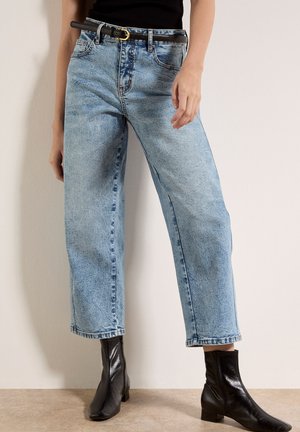 Donna che indossa jeans a gamba larga a vita alta azzurri con una cintura nera e stivali alla caviglia in pelle nera, in piedi contro un muro beige.