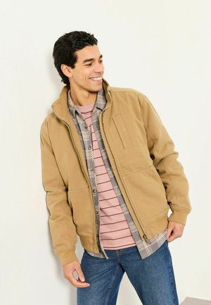 Braune Jacke mit Reißverschluss und hohem Kragen, getragen über einem karierten Hemd mit Knöpfen und einem gestreiften T-Shirt; verfügt über Taschen und eine lockere Passform.