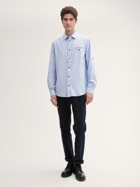 Chemise boutonnée à manches longues bleu clair avec une poche poitrine, boutons foncés, portée avec un pantalon marine et des chaussures noires à lacets. Texture lisse.
