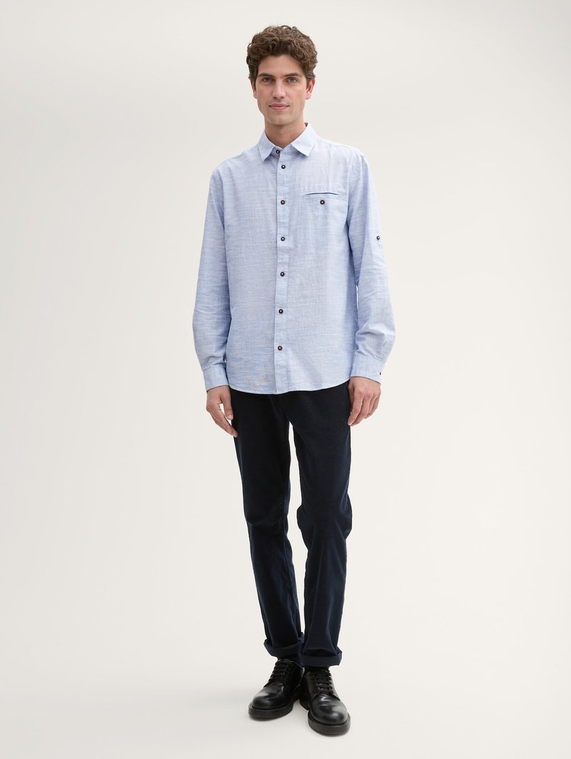 Chemise boutonnée à manches longues bleu clair avec une poche poitrine, boutons foncés, portée avec un pantalon marine et des chaussures noires à lacets. Texture lisse.