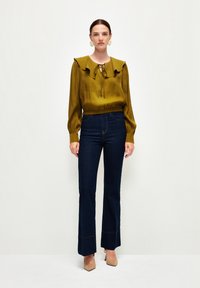 Olivengrøn bluse med flæser ved kraven og bindedetalje, parret med mørkeblå flagrende jeans. Glat tekstur, blanding af afslappede og sofistikerede elementer.