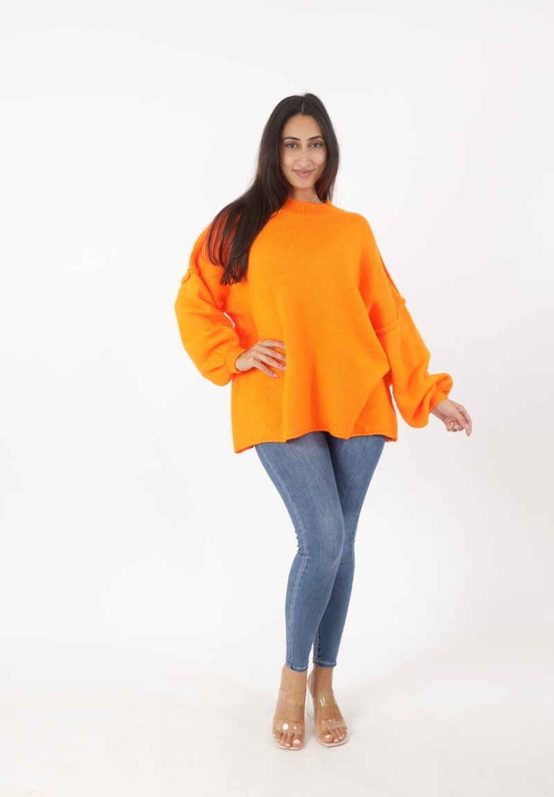 Helles orange Übergröße-Pullover mit geripptem Halsausschnitt und voluminösen Ärmeln, kombiniert mit enganliegenden blauen Jeans und sandalen mit transparentem Absatz.
