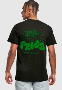 Mustan puuvillapaidan, jossa on vihreät grafiikat, takana ranskalaisia perunoita sisältävä astia ja teksti "FRIES CRISP VIBES ONLY".